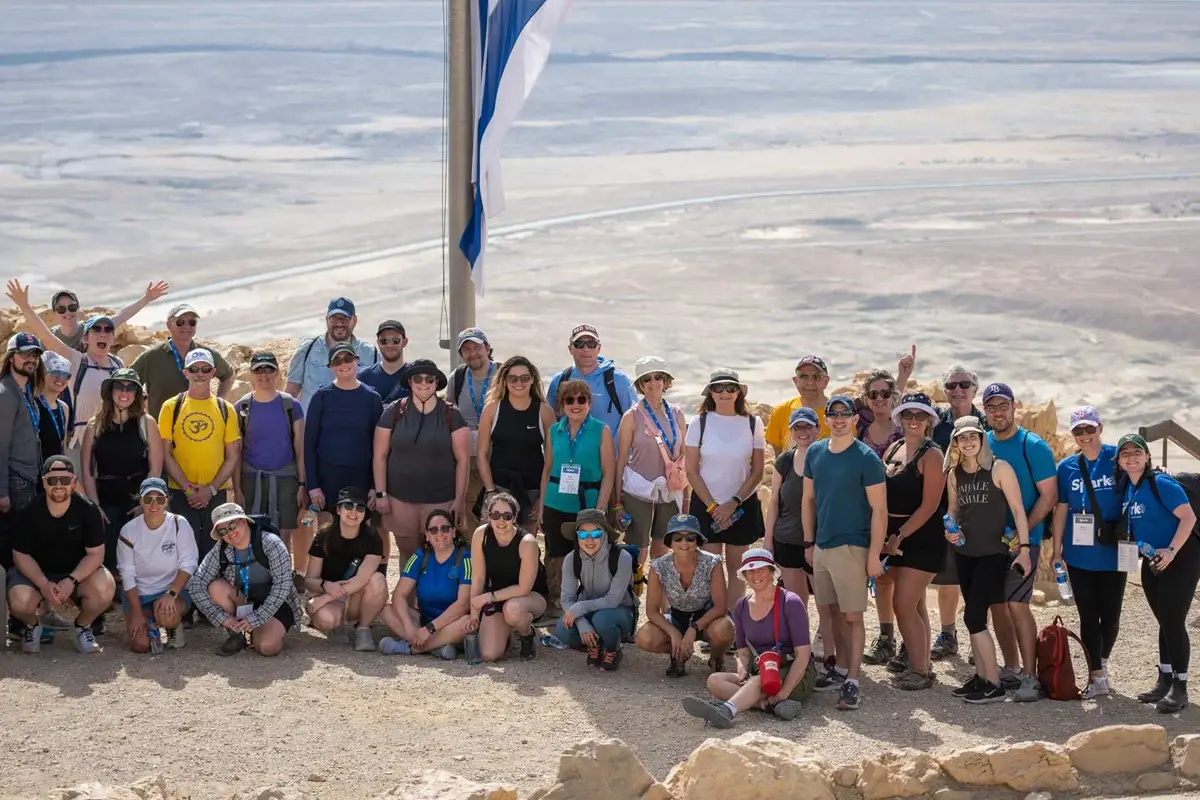 Young adult jewish life boston: A large group of young adults pose together during a community travel experience in a desert landscape beside an Israeli flag.