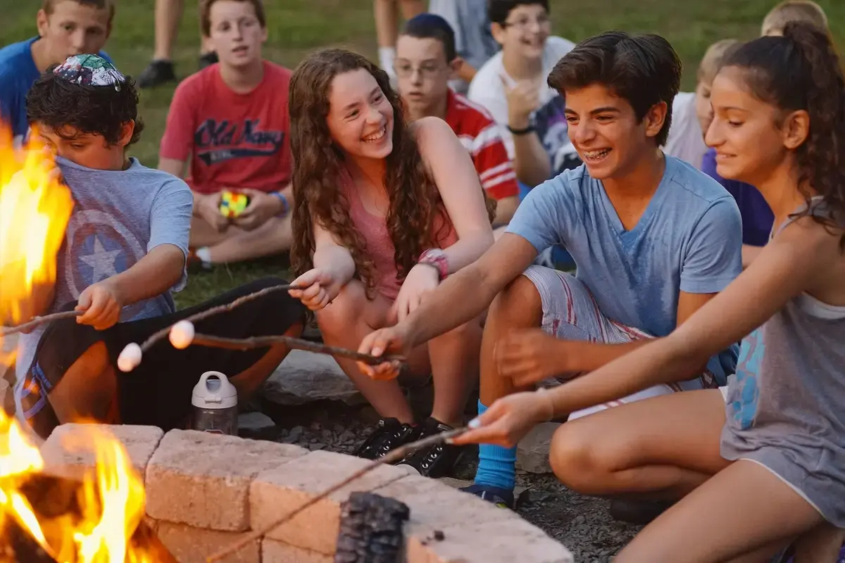 Jewish summer camp teens roasting marshmallows together around a campfire.