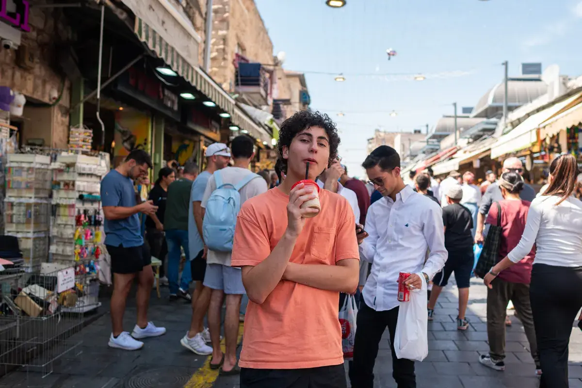 Israel teen travel boston: teen enjoying a drink while exploring a vibrant outdoor market in Israel, surrounded by shops, local culture, and bustling street life.